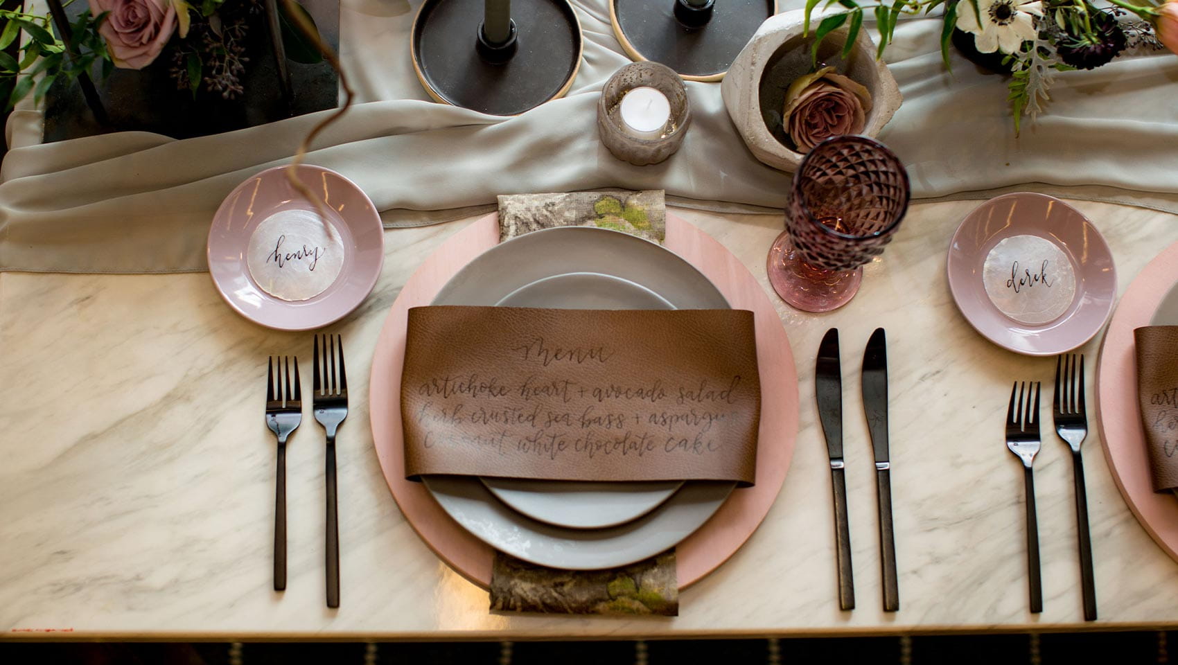 Elegant wedding table in Denver with name plates, a menu on a brown napkin, black utensils, pink plates, a purple glass, flowers, and candles.