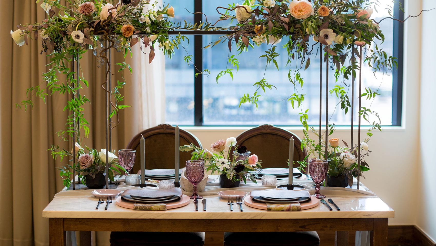 At Limelight Denver, an elegantly set dining table with floral arrangements and candles sits in a bright room with large windows and beige curtains.