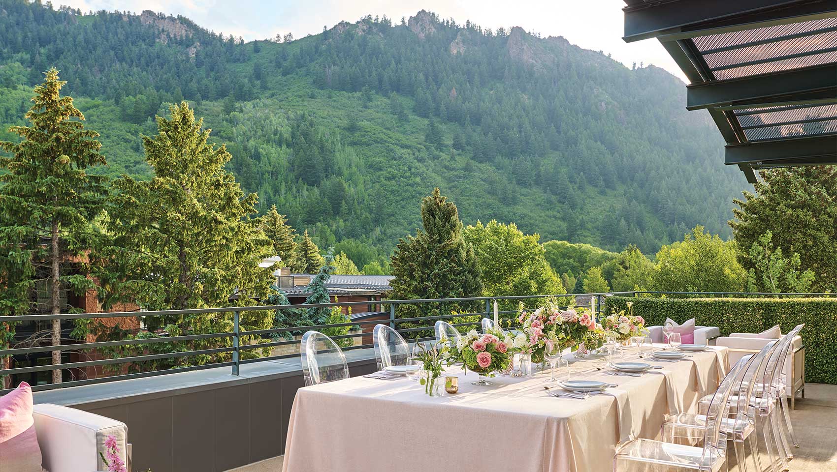 A table set for an outdoor meal with floral arrangements and transparent chairs on a terrace, surrounded by lush trees and mountains at Limelight Aspen.