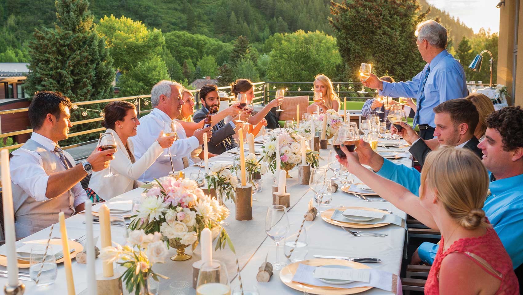 Guests toast around a candlelit, flower-adorned table on a patio in Aspen, surrounded by lush greenery and a mountainous backdrop.