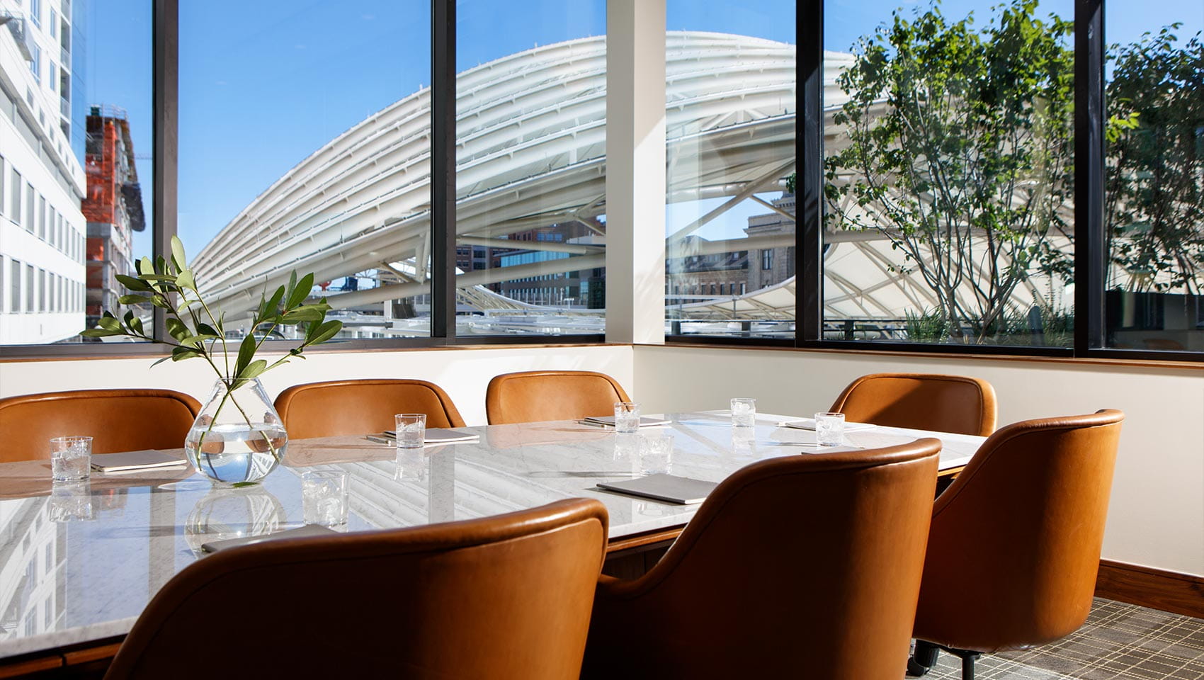 A marble conference table with brown leather chairs surrounds a glass vase holding green leaves. The room is well-lit by large windows revealing an architectural structure and trees outside.