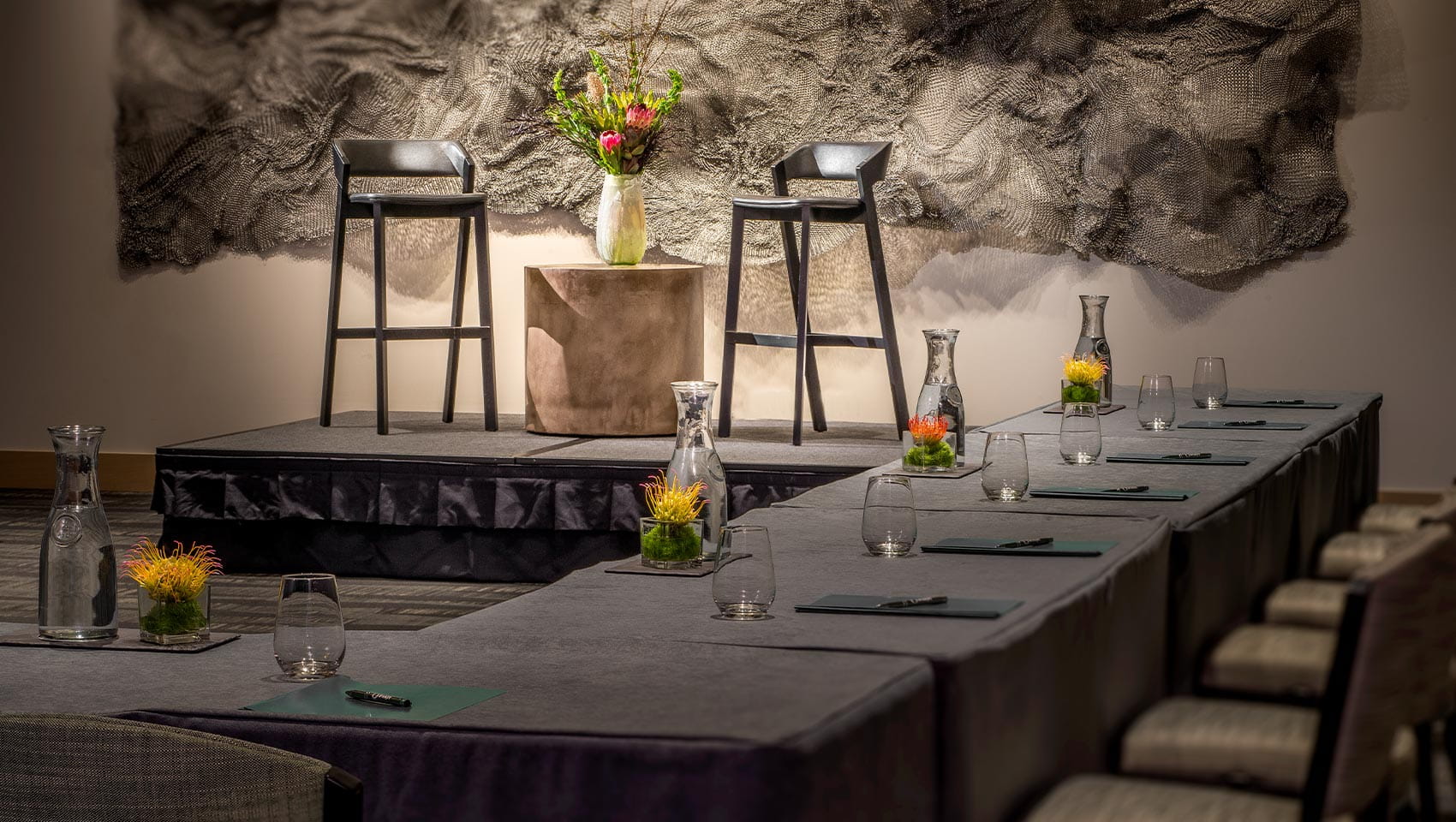 A small stage with two high chairs and a flower vase, surrounded by meeting tables with water carafes, glasses, notepads, and pens in a Limelight Ketchum conference room.