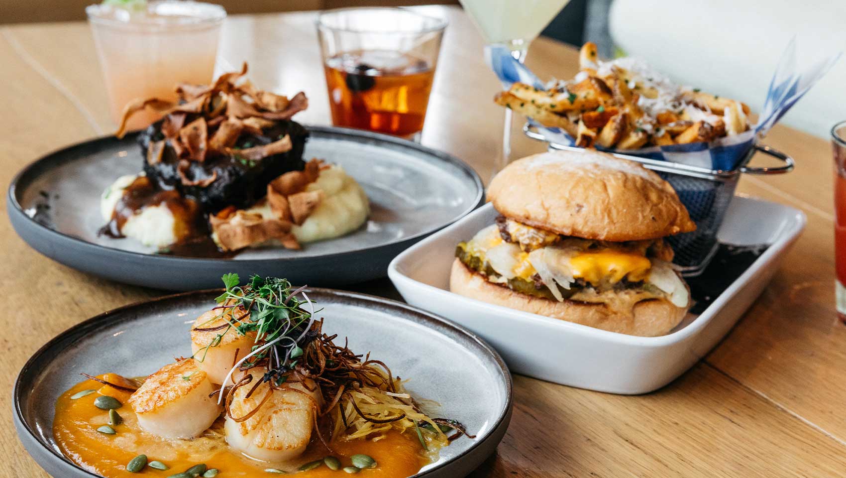 A table set with gourmet plates featuring seared scallops, a burger, a meat dish with mashed potatoes, and a basket of seasoned fries, accompanied by assorted beverages.