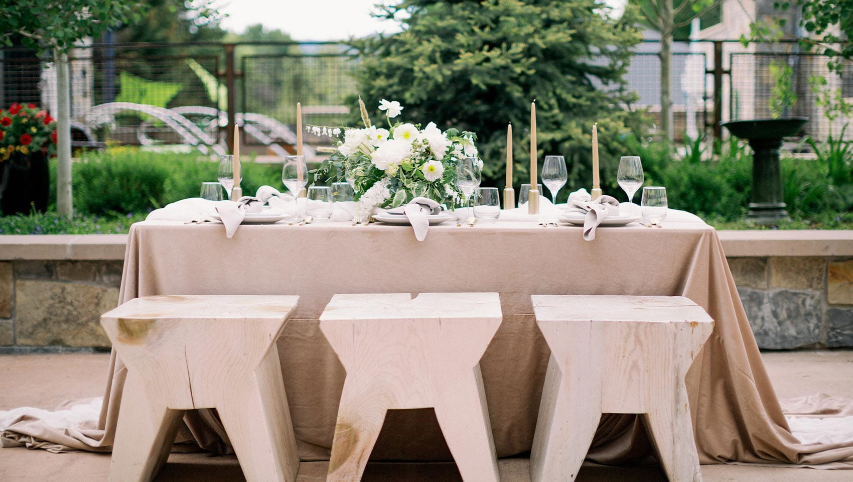 Looking at our Ketchum wedding venues, a table adorned with flowers, candles, and glassware, surrounded by wooden stools, is set in an outdoor garden with greenery and a stone wall.