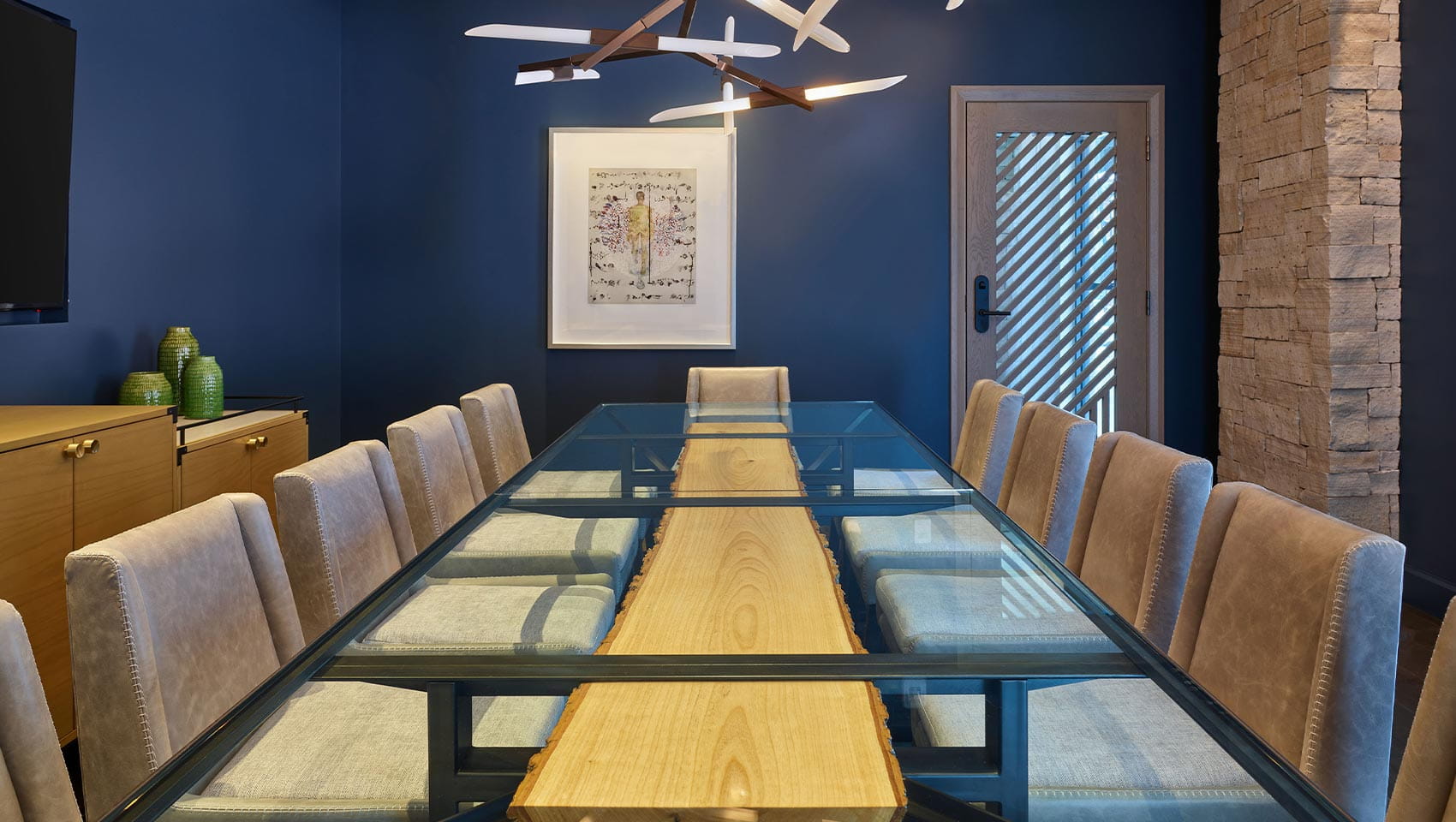 A long glass-top table with beige chairs under a modern chandelier in a blue-walled Limelight Snowmass conference room, with a wooden sideboard and artwork.