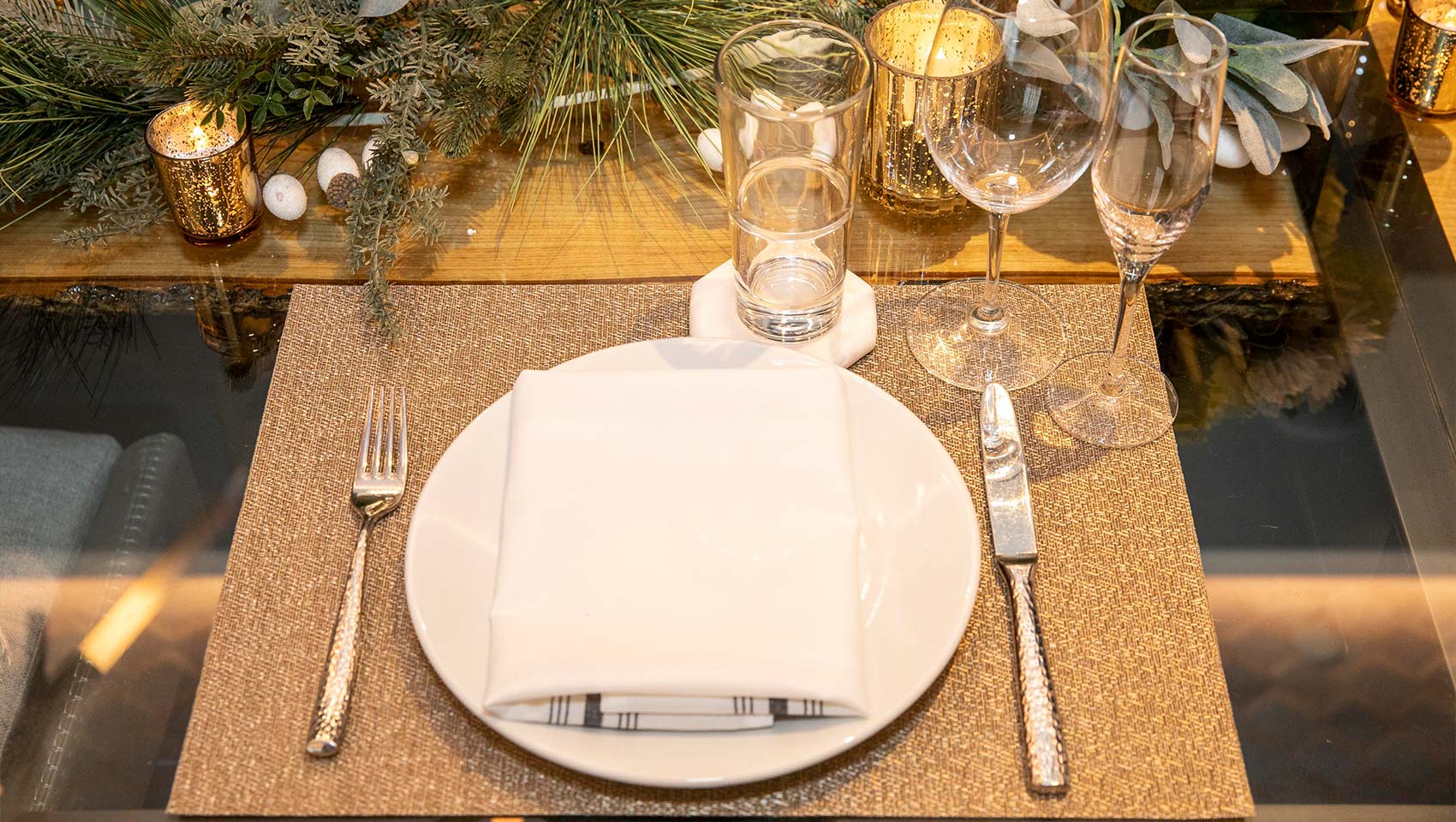 A table setting with a white plate, folded napkin, cutlery, glasses, and candles, surrounded by greenery, perfect for Snowmass weddings.