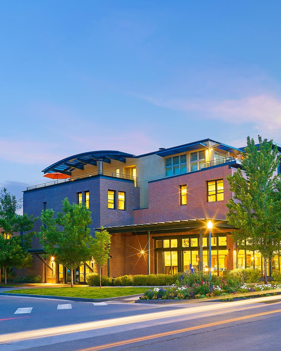 Limeight Aspen is a modern brick building with large windows is illuminated at dusk, featuring gardens, streetlights, and a crosswalk in front.