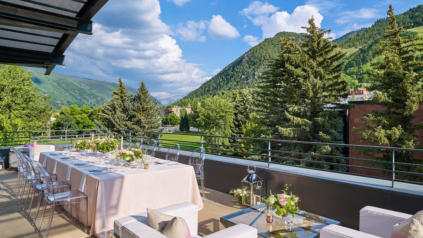 A pink tablecloth with floral arrangements on a terrace overlooking green trees and mountains, with transparent chairs, perfect for Aspen weddings.