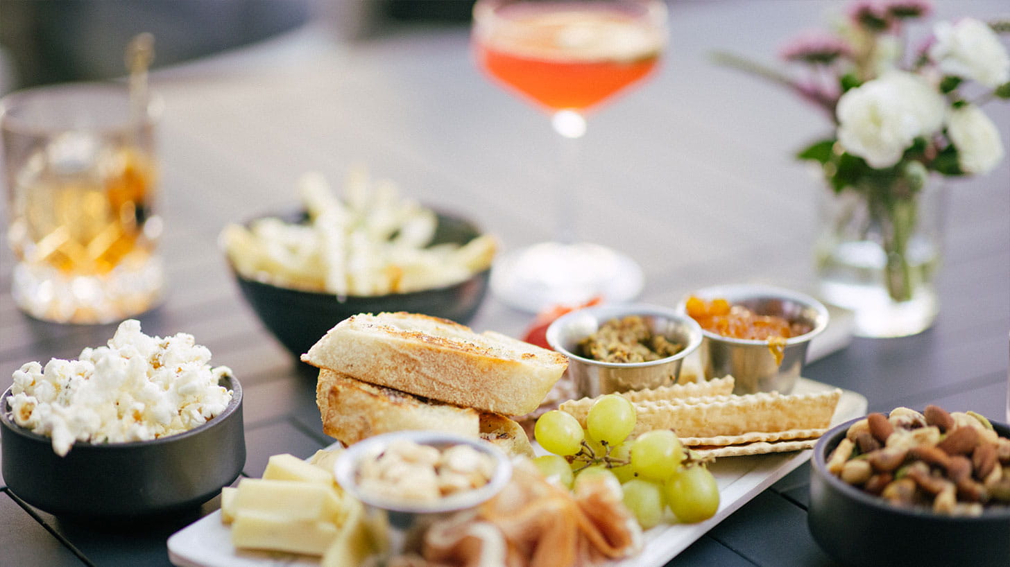 Food and drinks on a table in Aspen include bread, cheese, grapes, nuts, popcorn, fries, appetizers, a cocktail, and whiskey, with flowers.