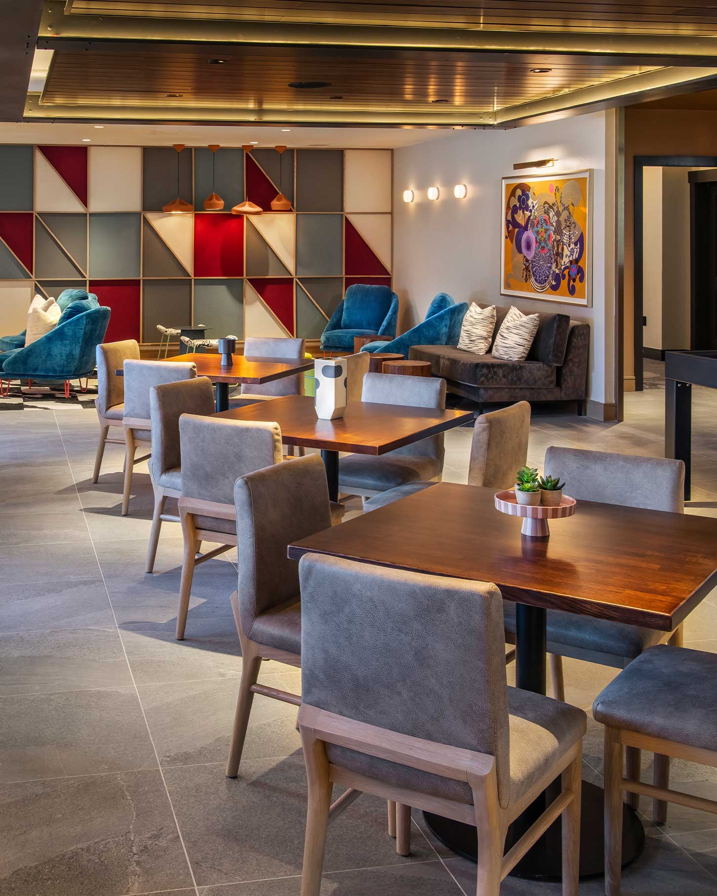 In the Limelight lounge in Aspen, wooden tables and padded chairs are arranged with blue armchairs, geometric wall decor, pendant lights, and contemporary artwork.