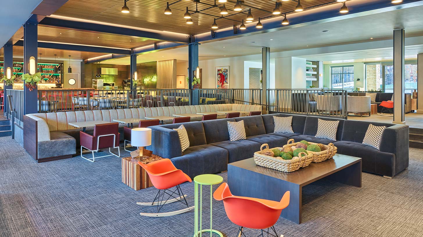 A large sectional sofa with patterned pillows in the Limelight Snowmass lounge area, with orange rocking chairs, tables, and a bar with vibrant decor.