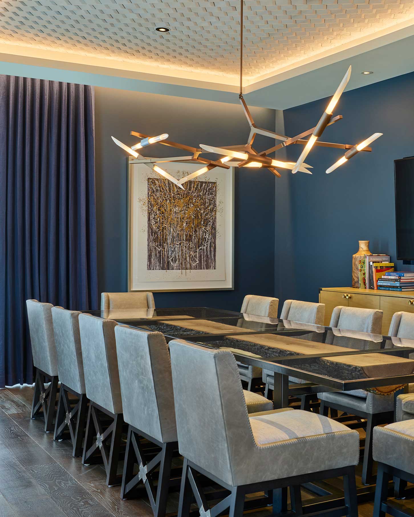 A dining table with gray chairs under a modern chandelier in a Limelight Snowmass room with blue walls, abstract art, and navy curtains.