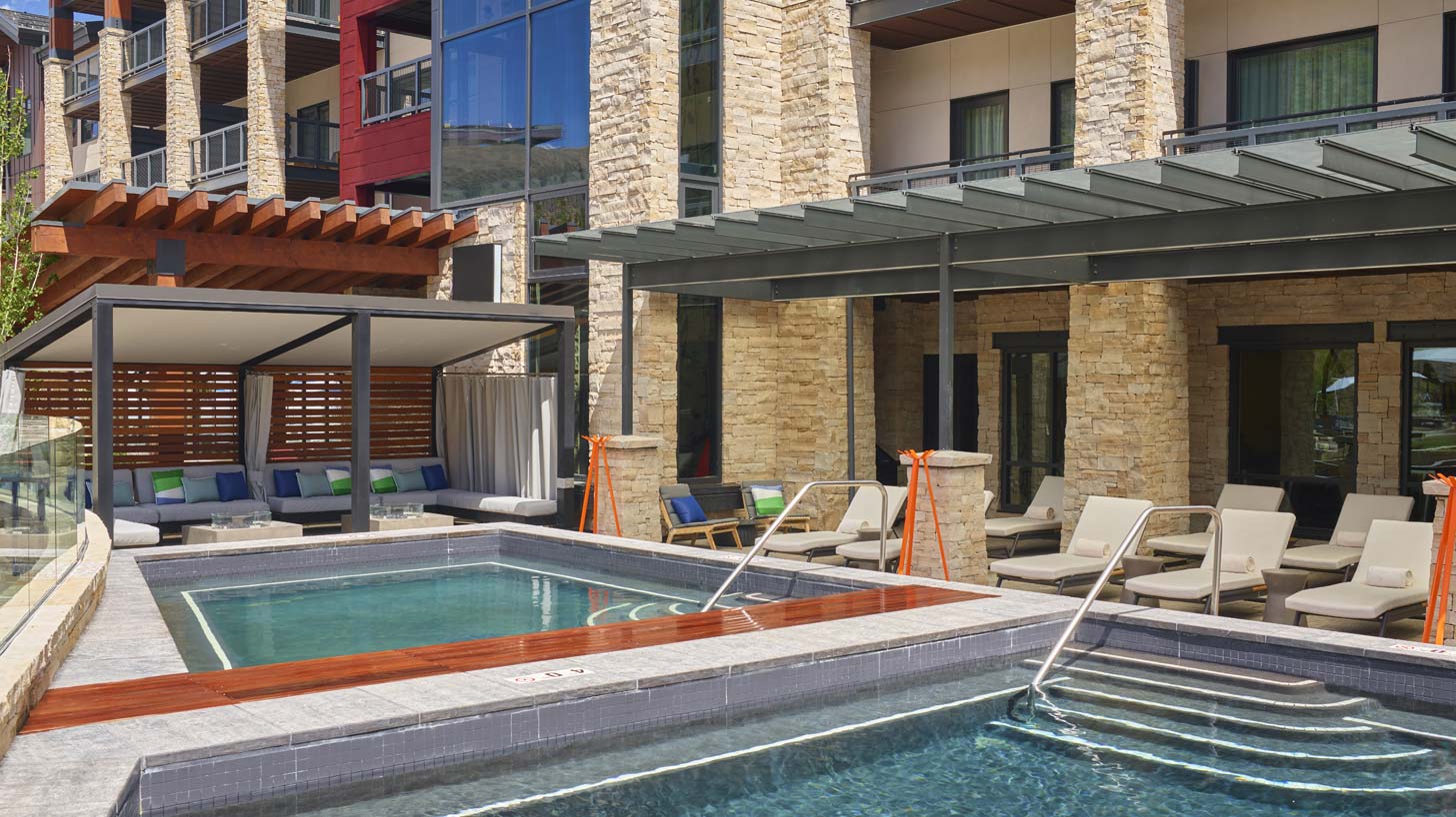 The Limelight Hotel pool area in Snowmass, Colorado features a hot tub, sun loungers, and shaded seating areas with cushions.