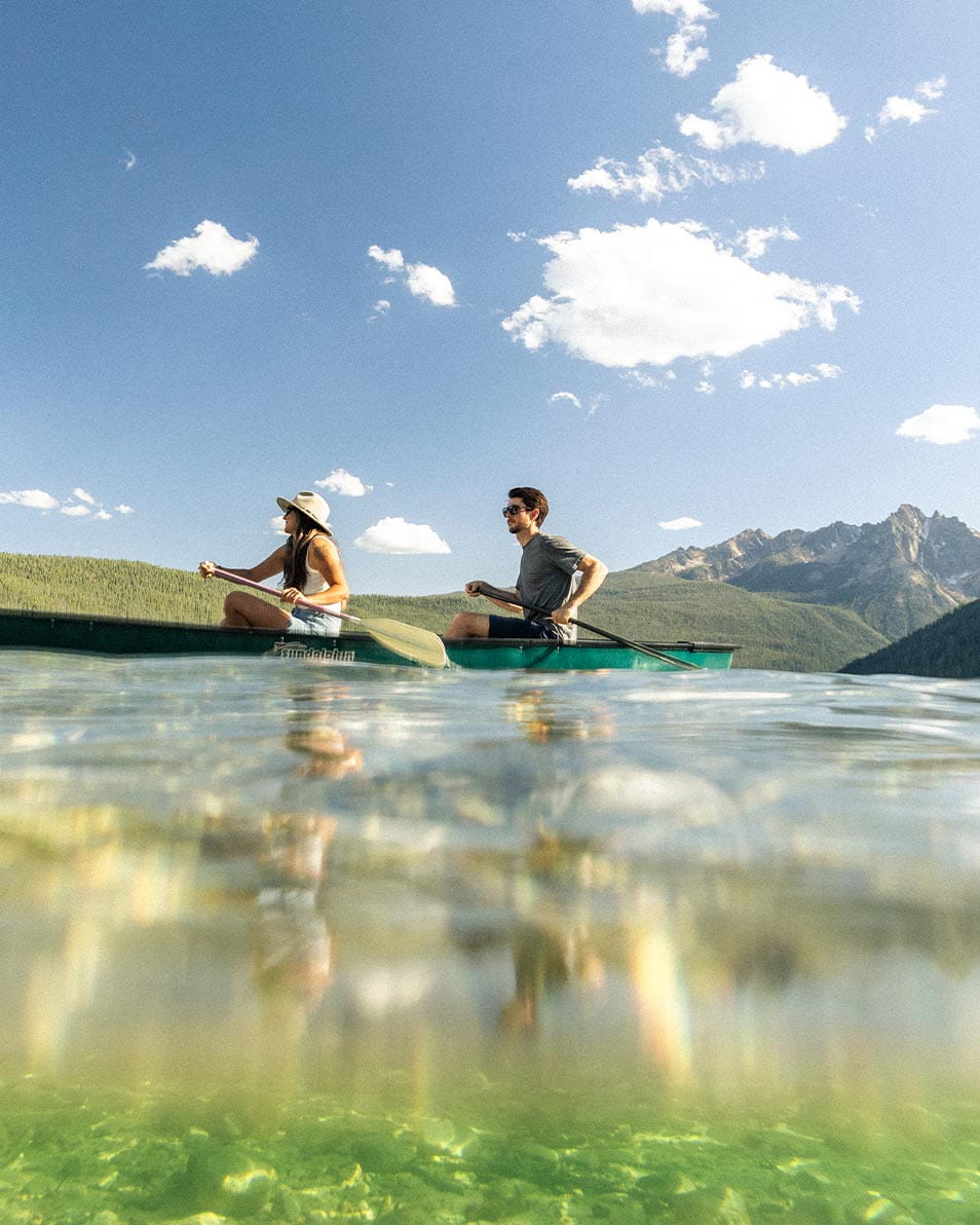 Two people paddle a canoe on a clear lake in Ketchum, with forested hills and mountains in the background under a bright, blue sky with scattered clouds.