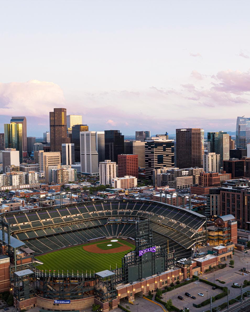 An empty Rockies stadium with a green field, surrounded by Denver's skyscrapers under a clear sky with scattered clouds after sunset.