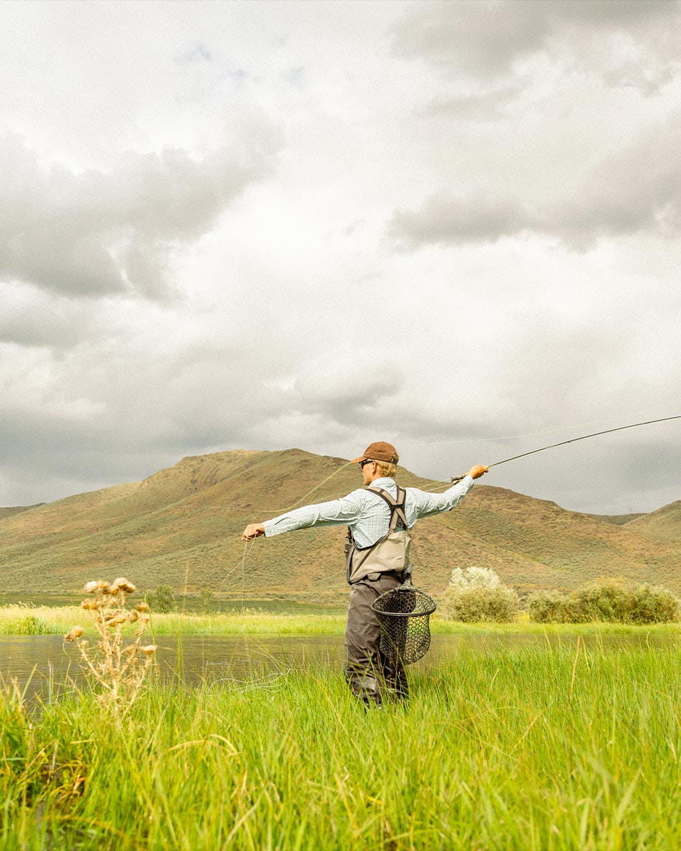In Ketchum, a person in wading gear casts a fishing line near a grassy riverside area, with rolling green hills and a cloudy sky in the backdrop.