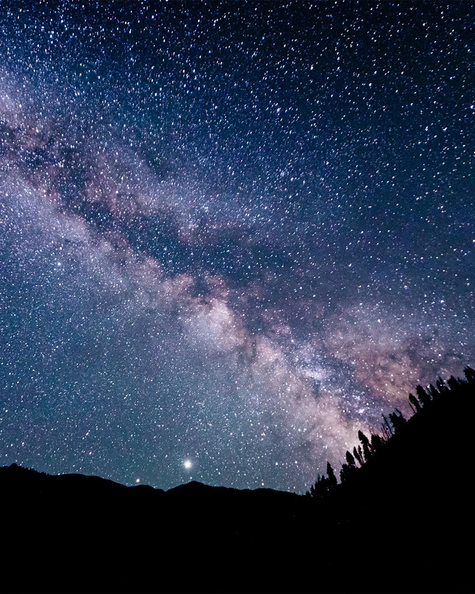 Stars and the Milky Way stretch across the night sky, with tree-lined mountains silhouetted below, visible from Ketchum.