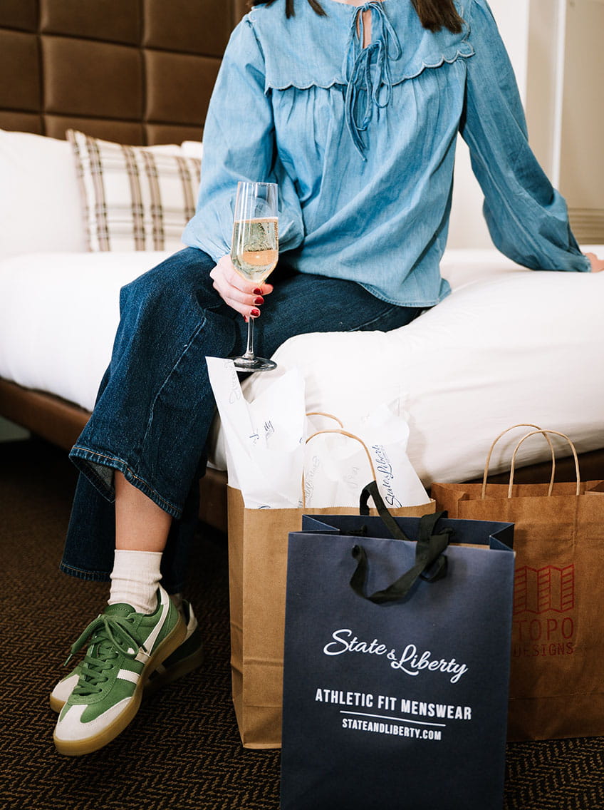 woman with champagne and shopping bags sitting on bed in guest room