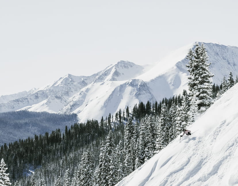 Skier skiing down a steep cliff next to trees and a mountain range in winter
