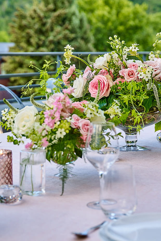 Floral arrangements with pink and white flowers adorn a table with glassware and plates on a terrace in Aspen, overlooking lush greenery.
