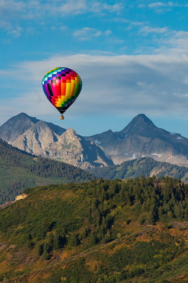 A multicolored hot air balloon floats in the sky above a forest and mountain range in Snowmass, CO under a partly cloudy blue sky.