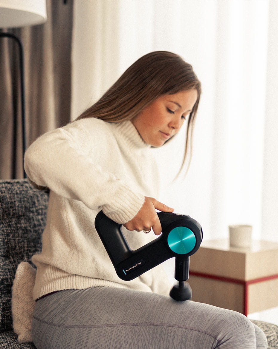 A person uses a massage gun on their thigh while sitting on a couch in a softly lit room with sheer curtains at the Limelight Aspen Hotel.