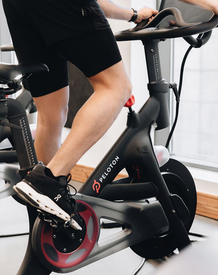 A person pedals a Peloton stationary bike in a modern workout room with large windows in Denver, wearing black athletic shorts and shoes.