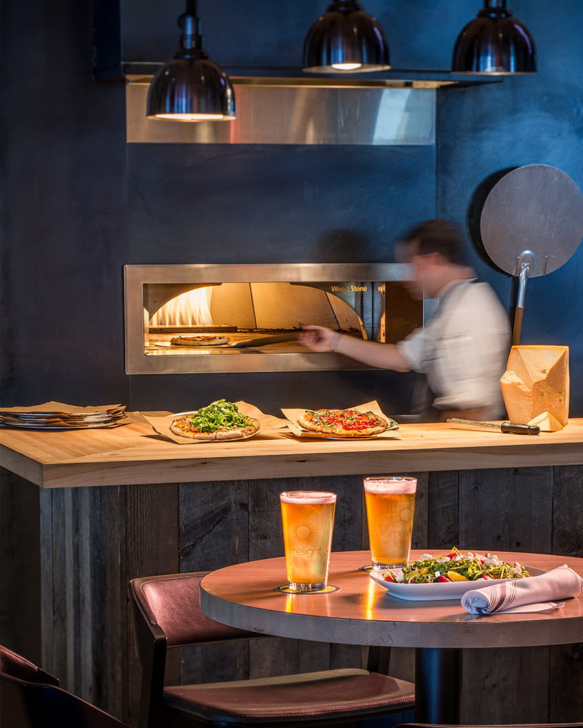 A chef at Limelight Hotel in Ketchum, Idago uses a pizza oven behind a wooden counter topped with pizzas.
