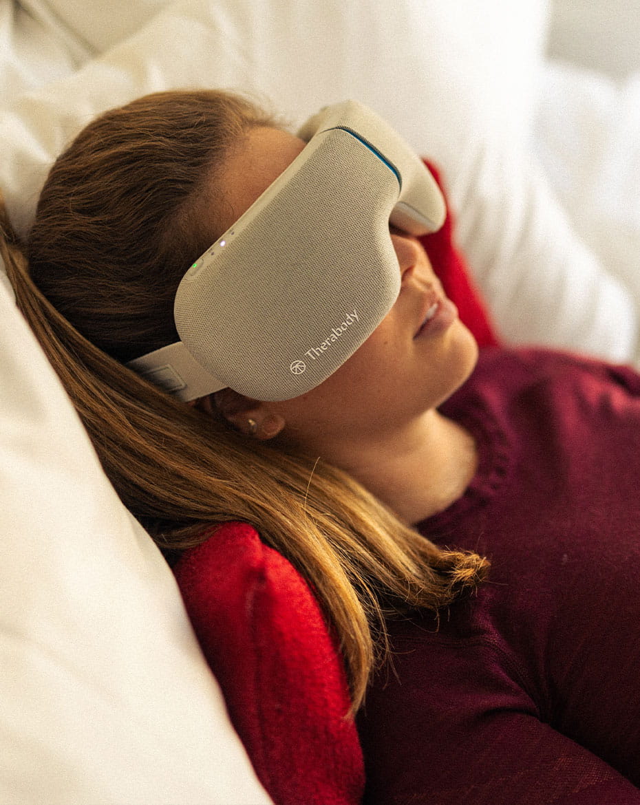 At Limelight Snowmass, a woman relaxes on a white pillow, wearing a gray "Therabody" eye massager and a maroon shirt, with her hair pulled back.