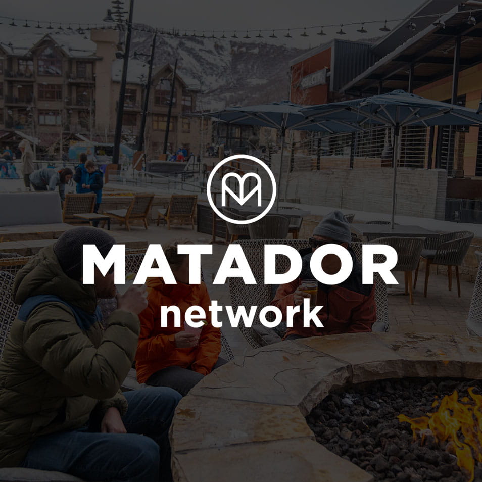 Three people in winter clothing sit around a fire pit. The background features snow-capped buildings and mountains. Text: "MATADOR network" with a circular logo containing an "M" above.