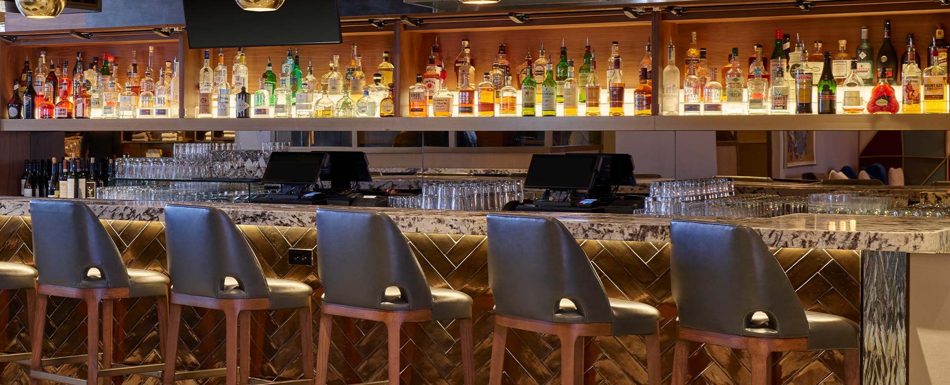 Chairs line a luxurious marble bar at the Limelight Aspen, with a well-lit shelf of various liquor bottles behind it in a modern, upscale bar setting.