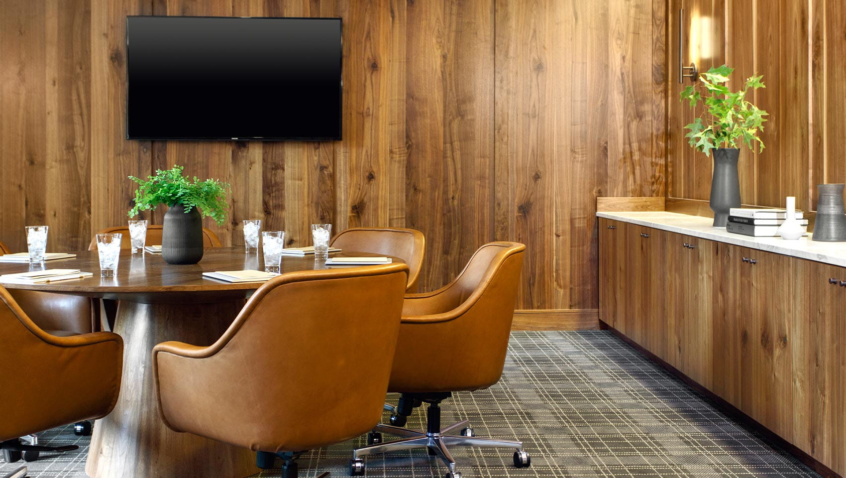 Round table set for a meeting, featuring a vase of greens in the center, surrounded by brown chairs in a wood-paneled room with a wall-mounted TV and sideboard.